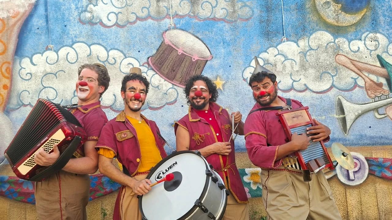 Quatro palhaços de nariz vermelho e figurino vinho, com sanfona, bumbo e tábua de lavar roupa, diante de um muro pintado com nuvens