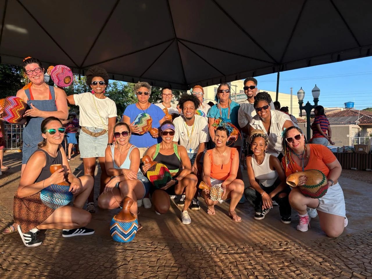Integrantes do grupo, sorridentes, posam sob uma tenda segurando xequerês coloridos