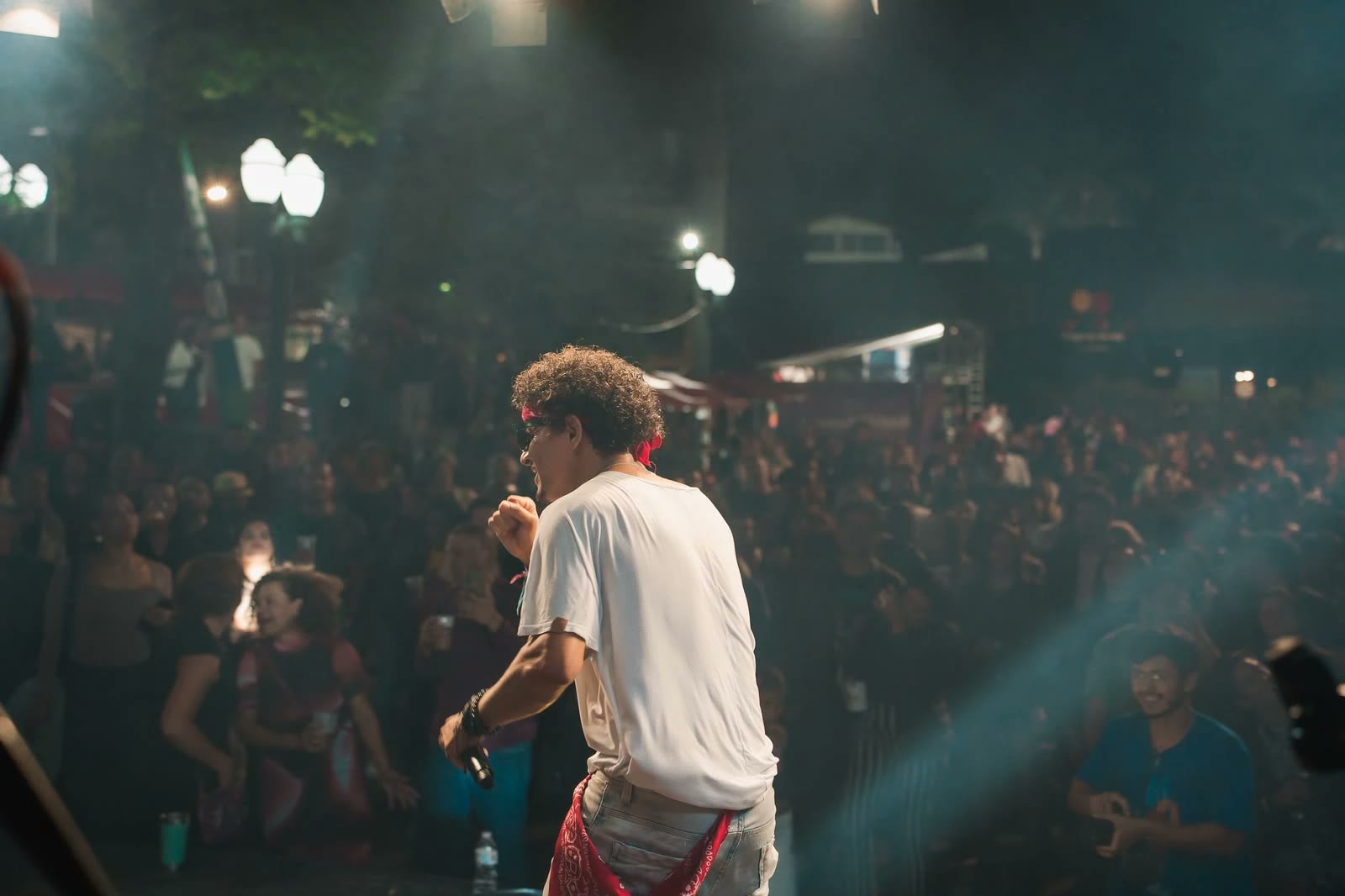 Artista de costas no palco, de camiseta branca e bandana vermelha, canta para a praça lotada à noite