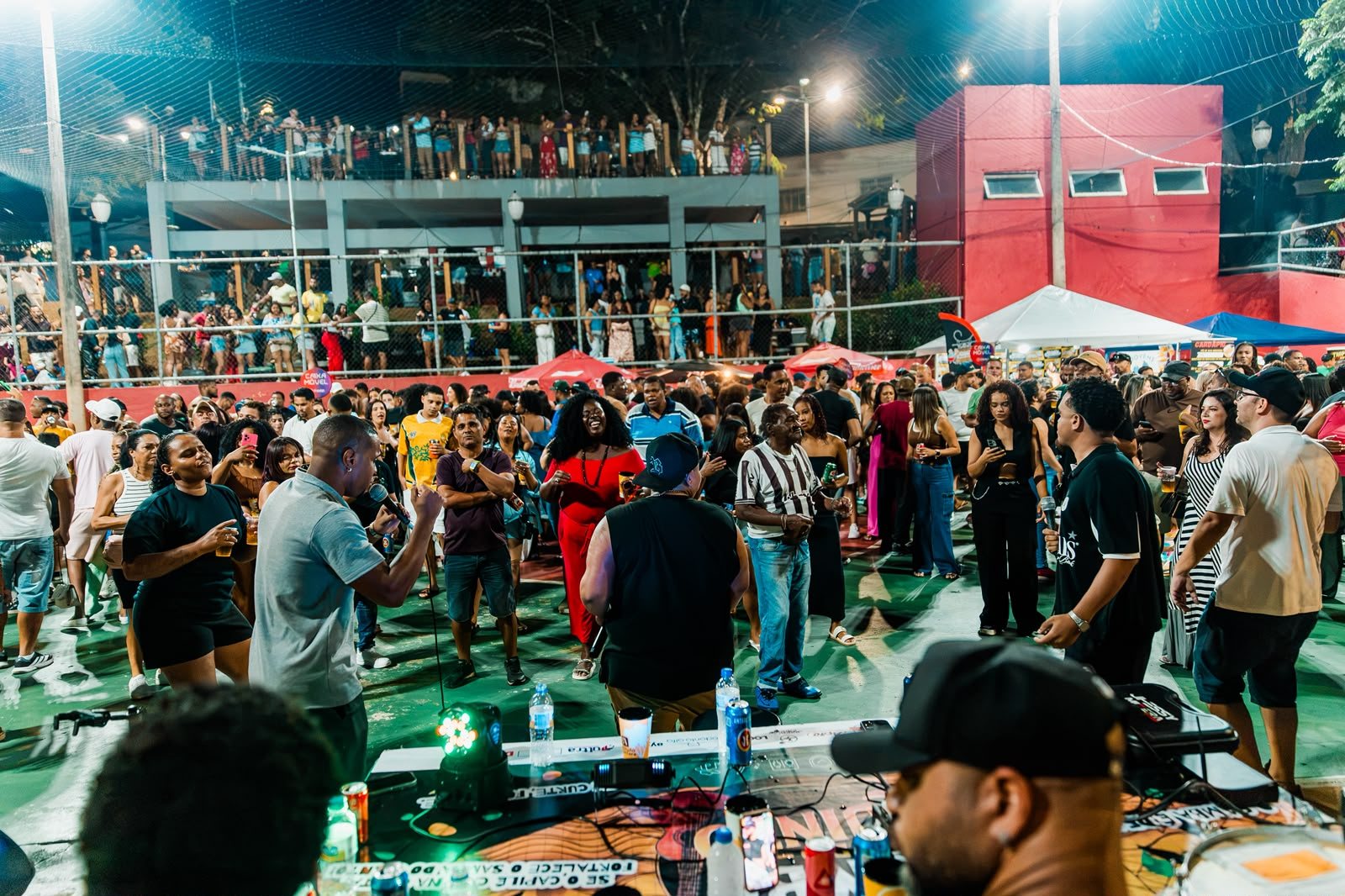 Cantores com microfone no meio do público, que dança na quadra; ao fundo, a arquibancada da praça lotada